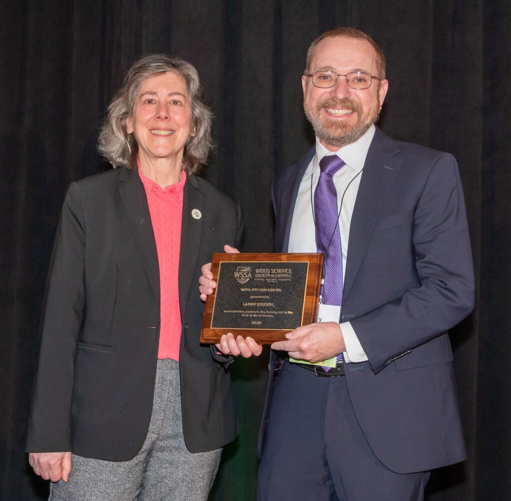 Two professionals at the Weed Science Society of America event, one presenting an award to the other, both smiling and dressed in formal attire.